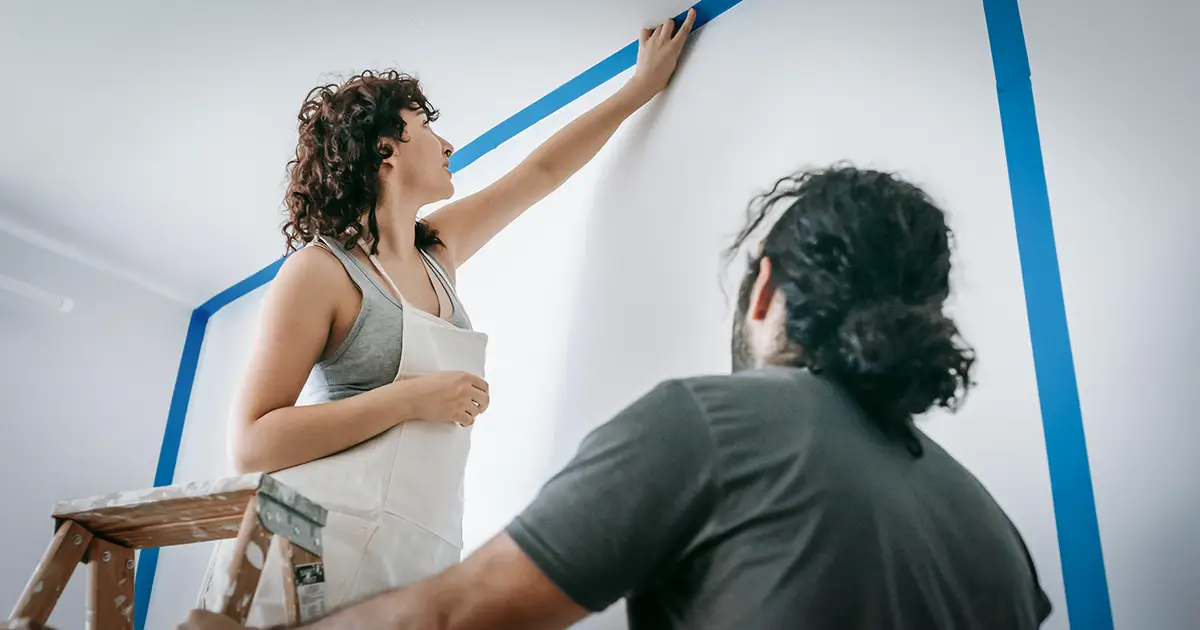 A woman stands on a ladder, putting up painter's tape for the upcoming remodel.